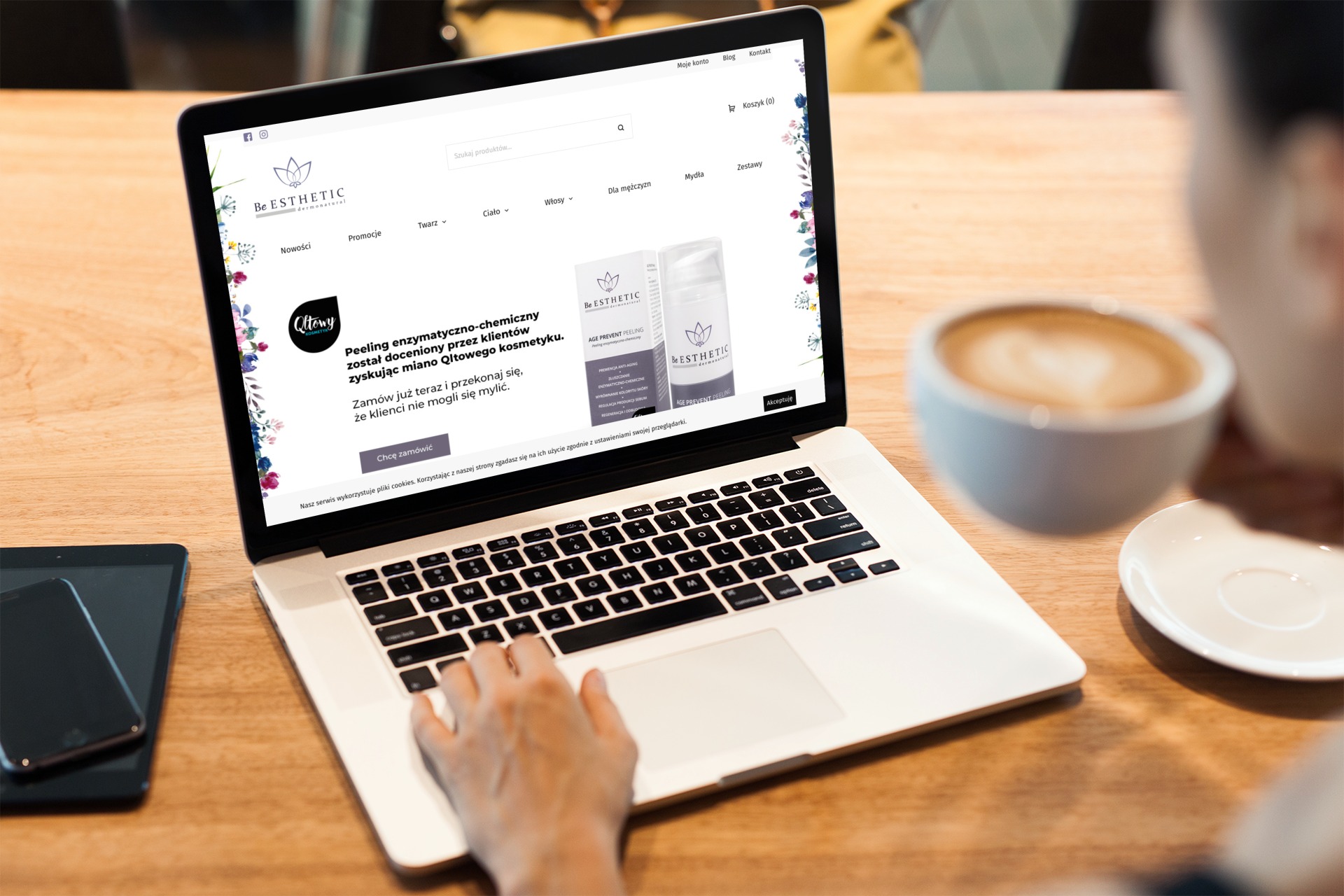 Osoba przegląda stronę internetową sklepu kosmetycznego 'Be Esthetic' na laptopie, obok widoczny tablet, smartfon i filiżanka kawy latte.