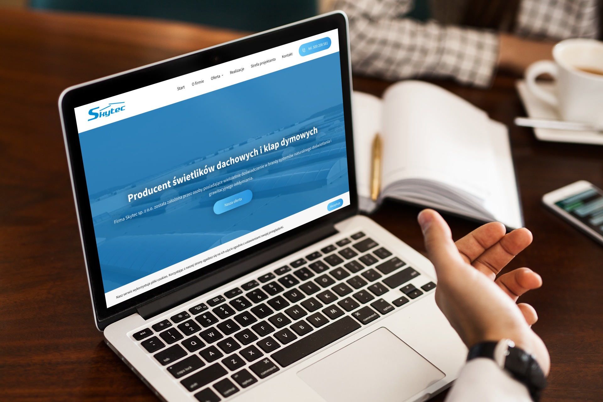 Laptop wyświetla stronę internetową firmy Skytec, producenta świetlików dachowych, z widoczną dłonią użytkownika na klawiaturze. W tle notatnik, kawa i smartfon.