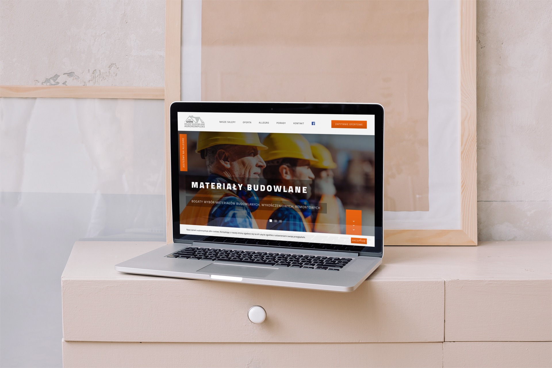 Laptop prezentuje stronę internetową firmy budowlanej z hasłem 'Materiały Budowlane', w tle widoczne ramy obrazów na tle ściany z delikatną fakturą.