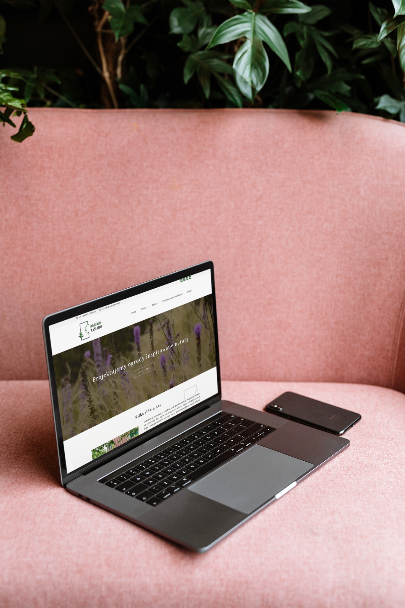 Laptop z wyświetloną stroną internetową o projektowaniu ogrodów, leżący na różowej kanapie obok smartfona, z zielonymi roślinami w tle.