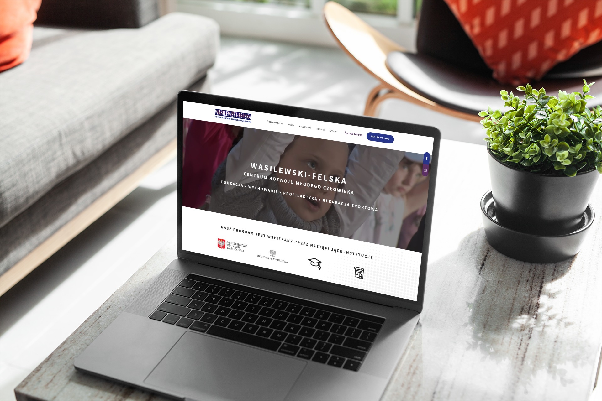 Laptop wyświetlający stronę internetową Wasilewski-Felska z ofertą edukacyjną i rozwojową dla dzieci, ustawiony na marmurowym blacie, obok doniczka z zieloną rośliną, w tle widoczne elementy...