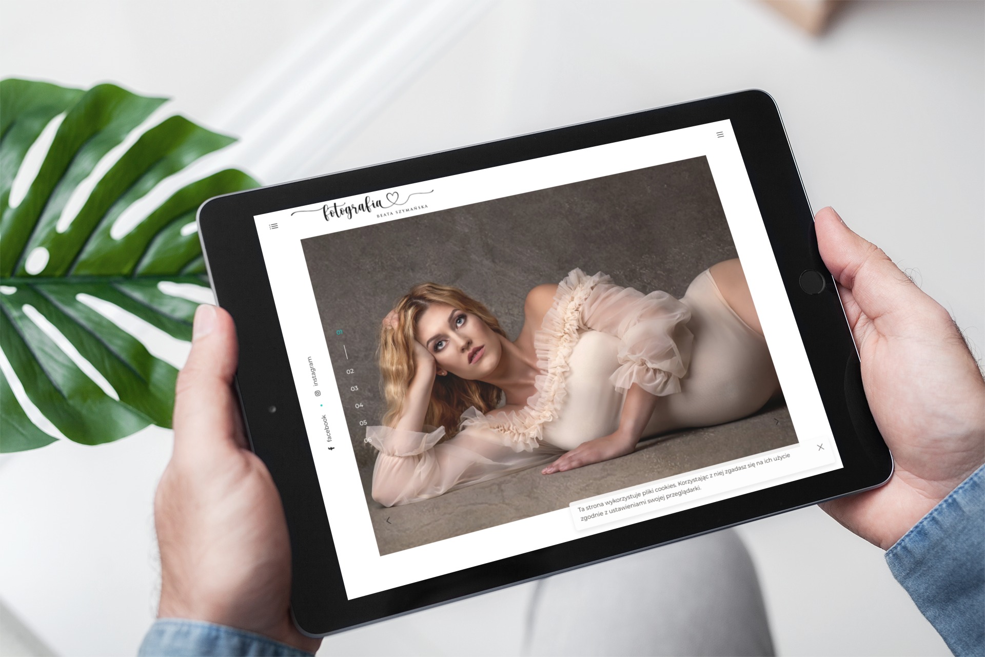 Mężczyzna trzyma tablet prezentujący stronę internetową z portretem kobiety w jasnej sukience, widoczne logo fotografa i menu strony, w tle liść monstery.