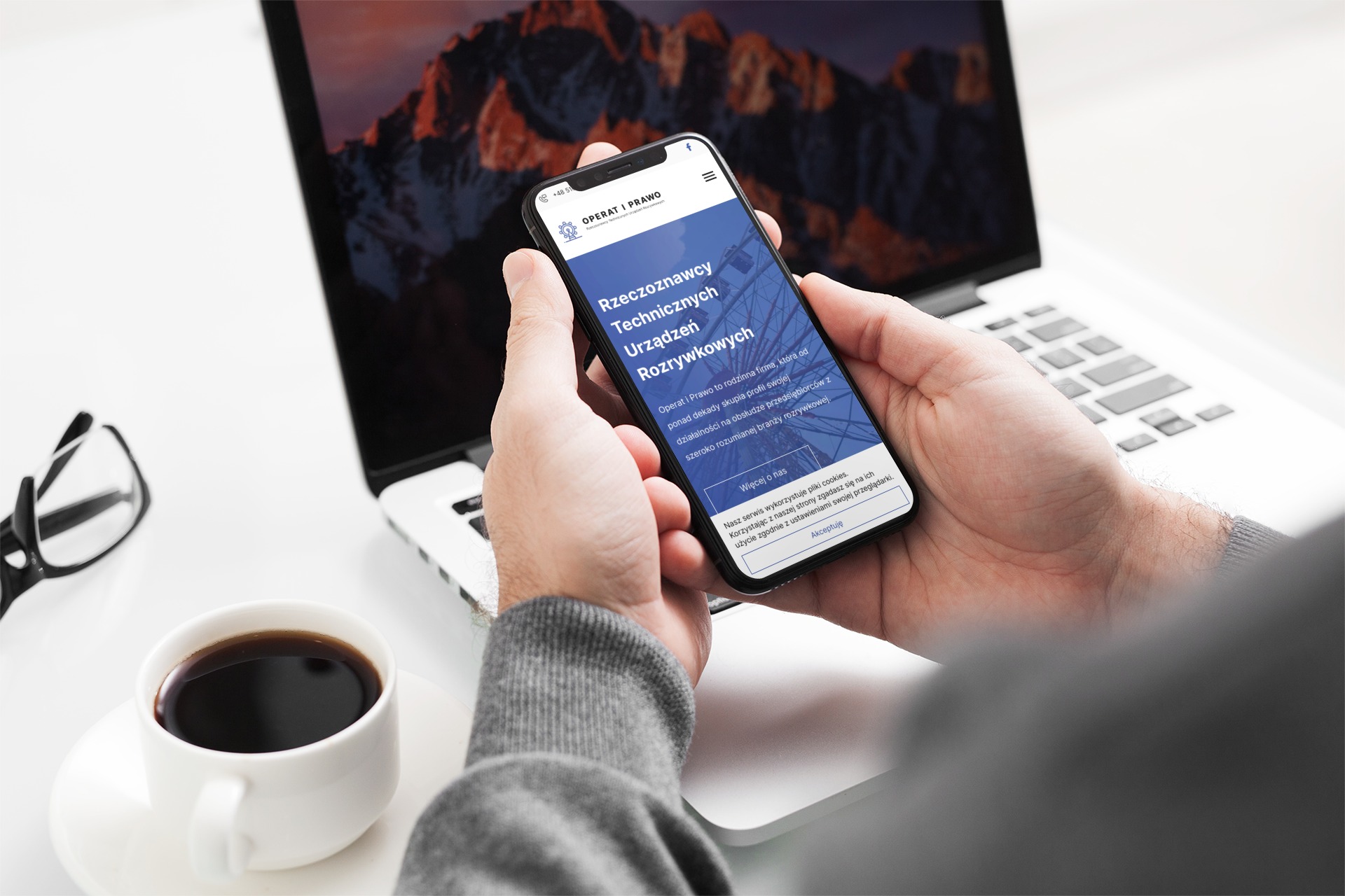 Osoba trzyma smartfon z wyświetloną stroną internetową Operat i Prawo na tle laptopa z wygaszaczem ekranu przedstawiającym góry, obok filiżanka kawy i okulary.