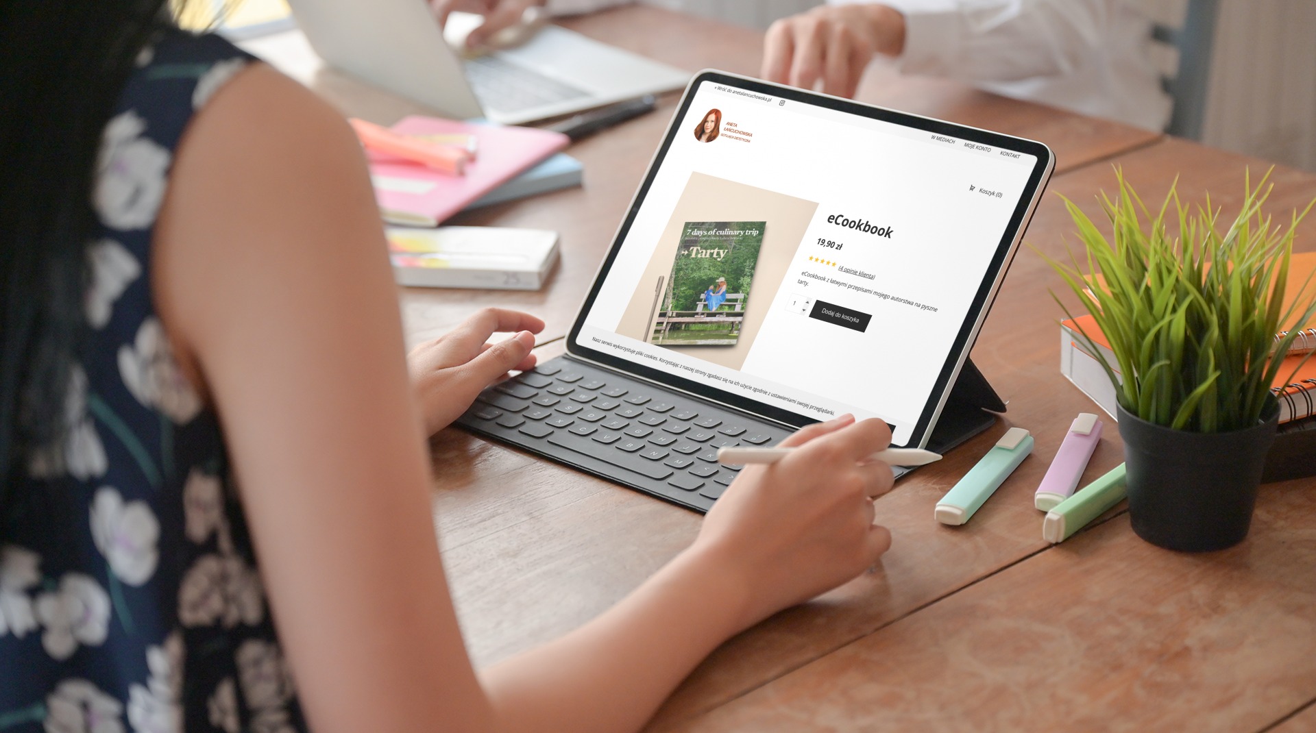 Osoba przegląda ofertę e-booka kulinarnego na tablecie z klawiaturą, używając rysika; na drewnianym biurku leżą zakreślacze i doniczka z zieloną rośliną.