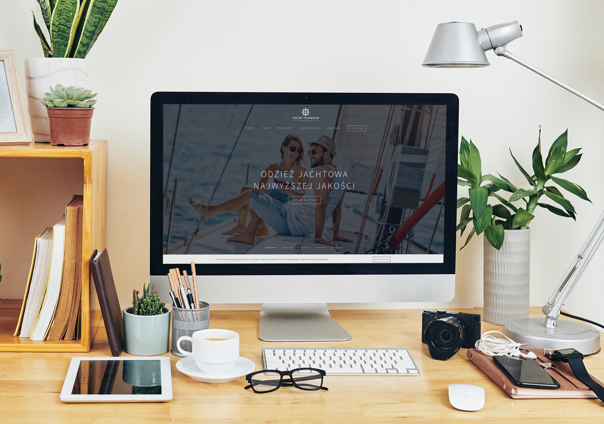 Monitor wyświetla stronę internetową z parą w odzieży jachtowej na jachcie, na biurku obok leżą okulary, klawiatura, tablet, aparat, telefon, słuchawki i kawa.