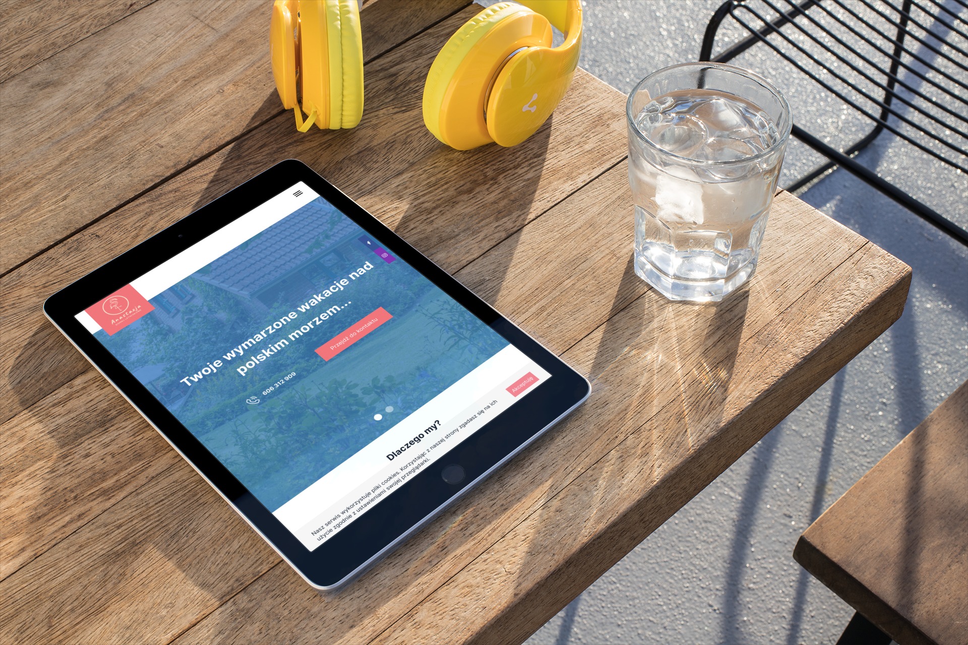 Tablet wyświetla stronę internetową z ofertą wakacji nad morzem, obok słuchawki i szklanka wody na drewnianym stole w słoneczny dzień.
