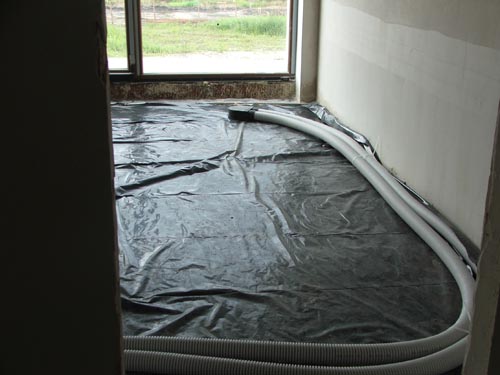 Przygotowanie podłoża pod system wentylacji z odzyskiem ciepła w surowym pomieszczeniu: czarna folia ochronna, białe rury wentylacyjne ułożone wzdłuż ściany, widok przez framugę drzwi.