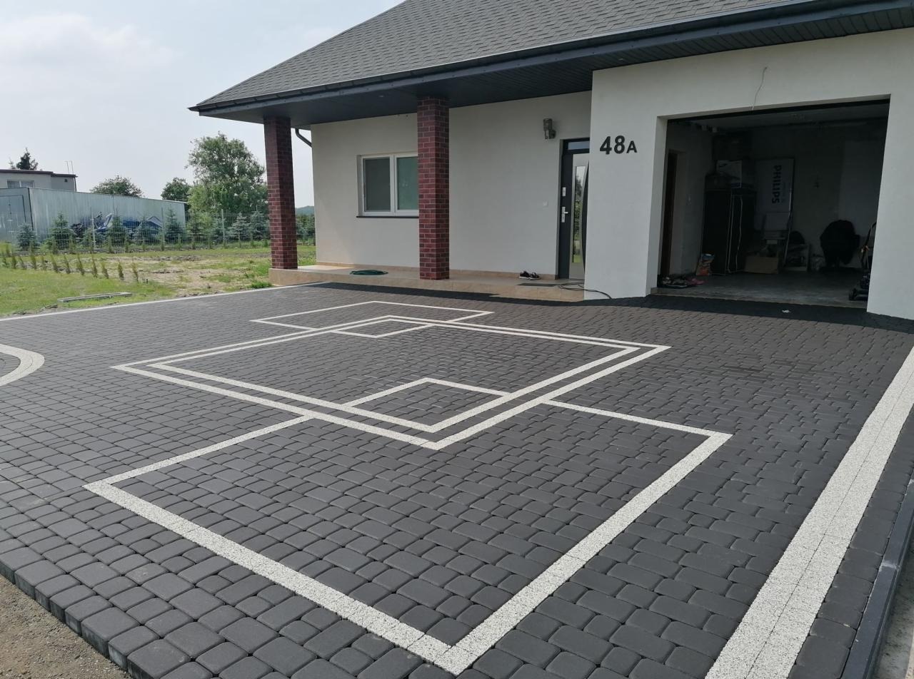 Nowoczesny podjazd z ciemnoszarej kostki brukowej z białymi, geometrycznymi wzorami, na tle domu z garażem i numerem 48A.