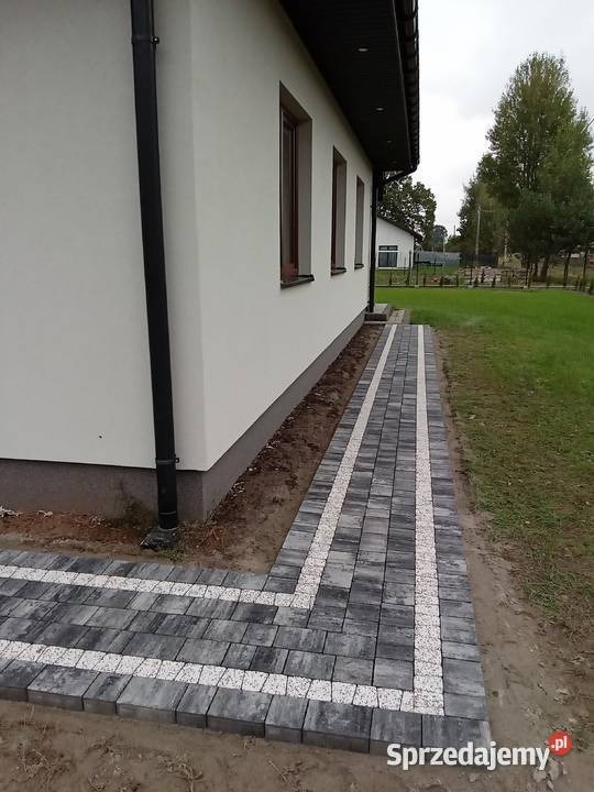 Szara kostka brukowa z białymi akcentami ułożona wzdłuż ściany budynku z rynną spustową, widok z boku, trawnik w tle.
