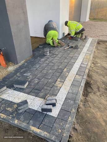 Dwóch pracowników w odblaskowych ubraniach układa ciemnoszarą kostkę brukową z jasnym, granitowym obramowaniem na podjeździe przed budynkiem.