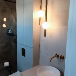 Nowoczesna łazienka z białą umywalką nablatową, złotą armaturą i wiszącymi lampami ze złotymi elementami, obok zabudowa szafkowa z ukrytą spłuczką WC, w tle kabina prysznicowa.