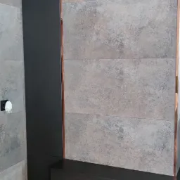 Nowoczesna ściana z szarych płytek imitujących kamień, obudowana czarną zabudową meblową z miedzianymi akcentami, tworząca spójny element dekoracyjny wnętrza.