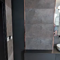 Nowoczesna łazienka z szarymi, teksturowanymi płytkami, czarnymi szafkami pod umywalką i miedzianymi akcentami, widoczny fragment lustra z odbiciem osoby.