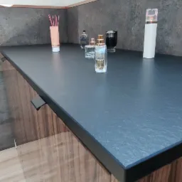 Ciemny blat kuchenny z różnymi flakonami perfum i dyfuzorem zapachowym, szafka z drewnopodobną okleiną.