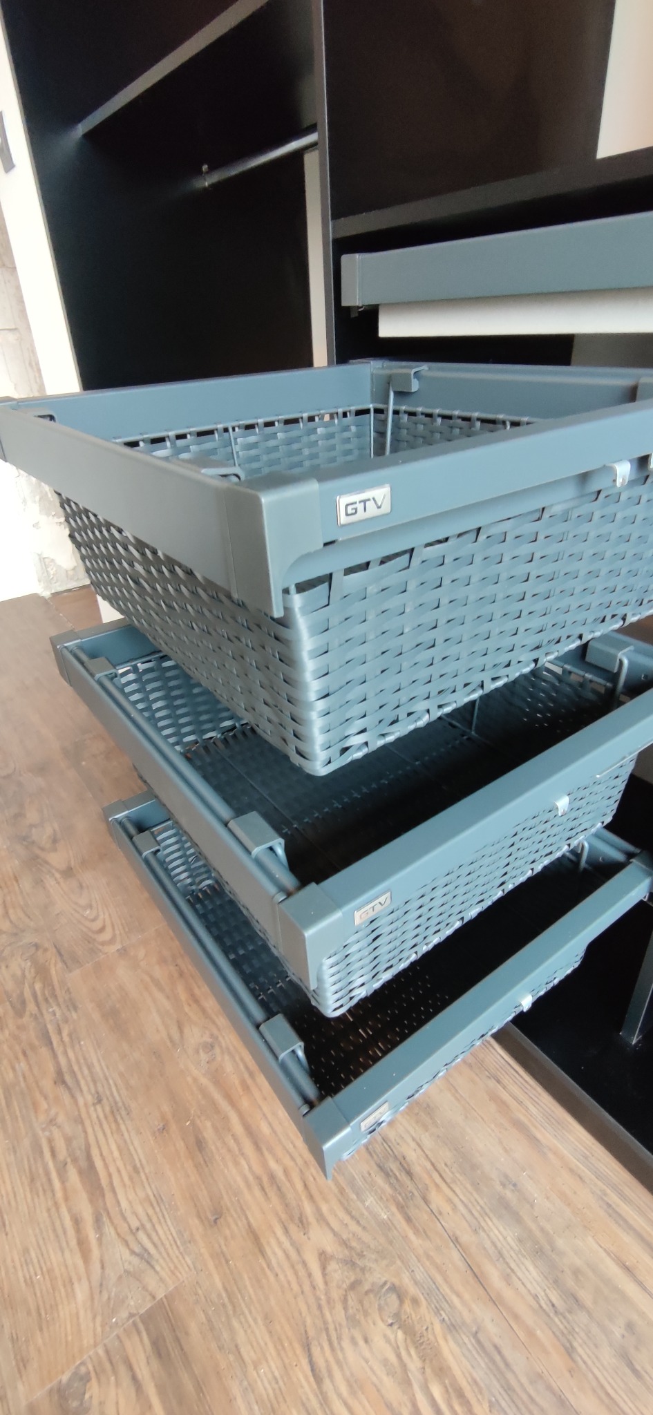 Wysuwane, szare kosze z plecionki w szafie, widoczne logo GTV na górnym koszu.
