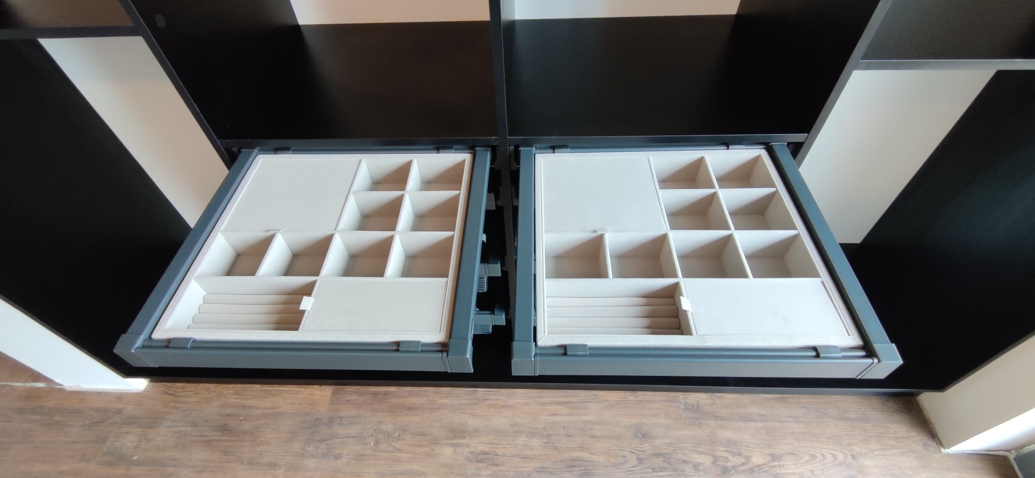 Dwie wysunięte szuflady z przegródkami, wyściełane jasnym materiałem, w czarno-białej szafie na drewnianej podłodze.