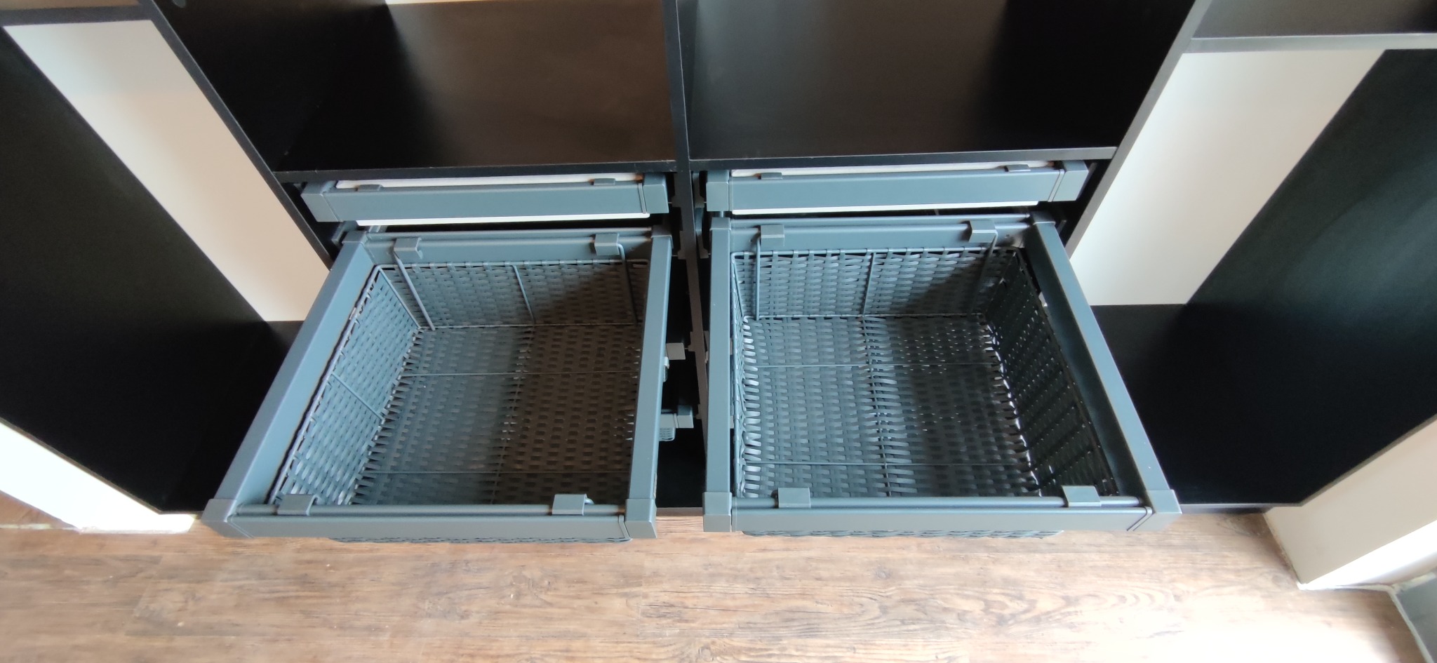 Dwie wysuwane, puste szuflady z metalowej siatki w szafie z czarnymi frontami, widok z góry na drewnianą podłogę.