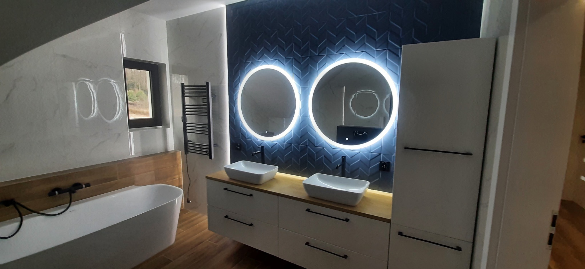 Nowoczesna łazienka z dwoma okrągłymi lustrami z podświetleniem LED, umywalkami nablatowymi na drewnianym blacie i ciemnymi płytkami chevron na ścianie.