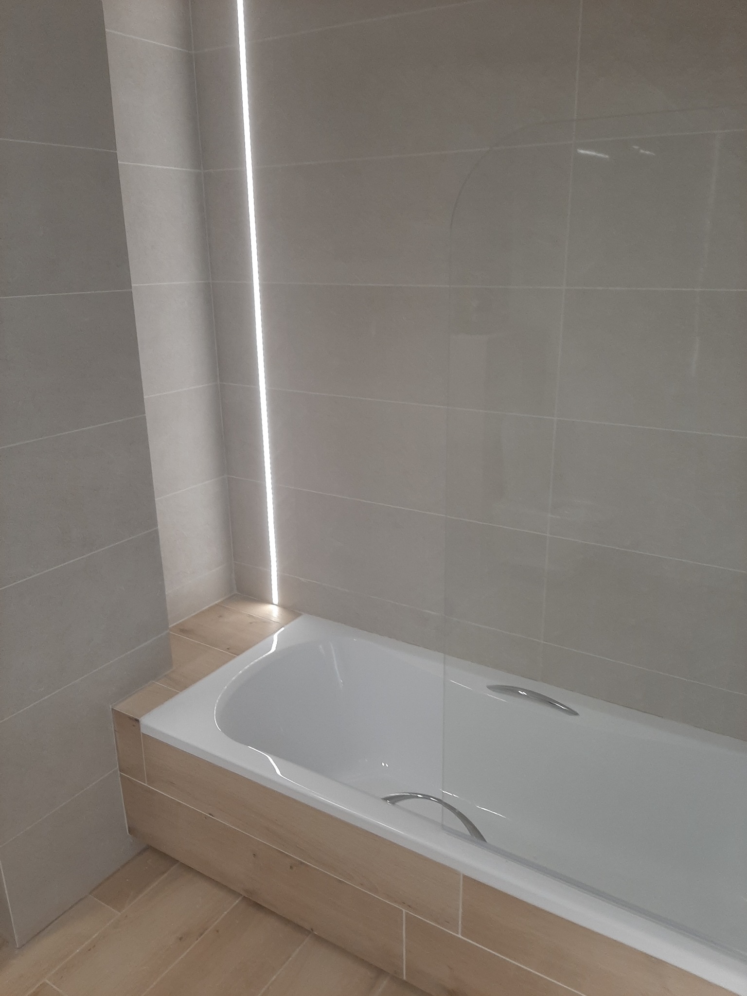 Biała wanna z chromowanymi uchwytami, obudowana panelami imitującymi drewno, jasnoszare płytki na ścianach z pionowym oświetleniem LED w narożniku, widok na fragment łazienki po remoncie.