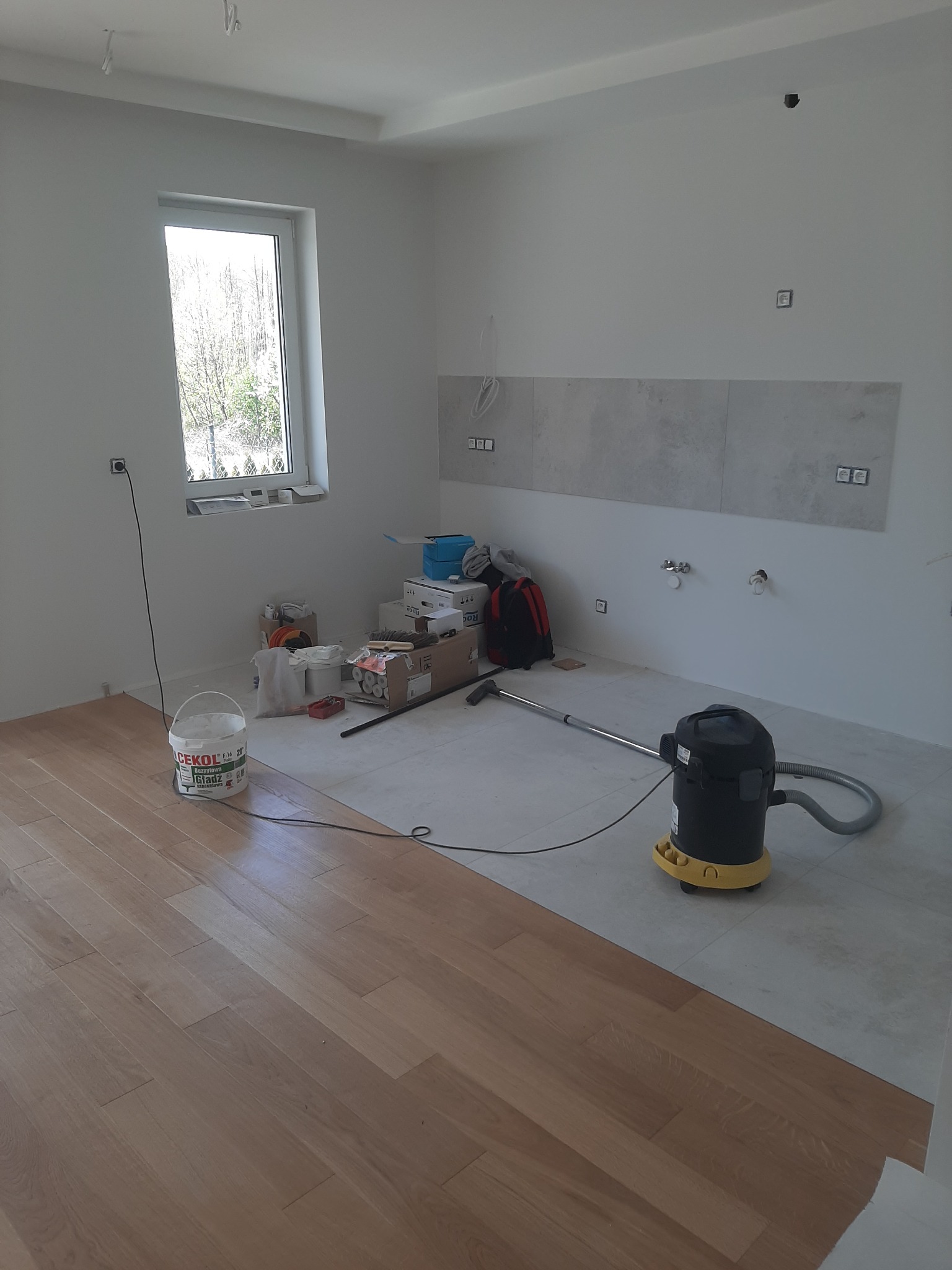 Fragment pokoju w trakcie prac wykończeniowych: położona drewniana podłoga kontrastuje z surową, szarą podłogą oraz częściowo wykończoną ścianą z szarymi płytkami, widoczny odkurzacz przemysłowy...
