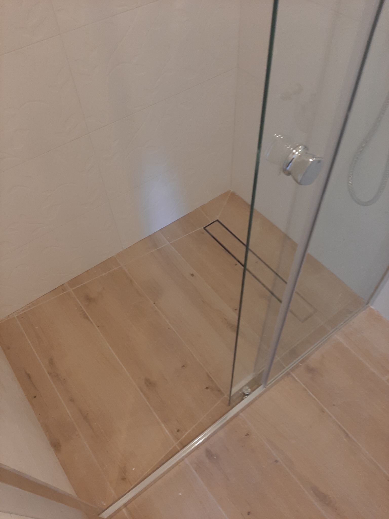 Nowoczesna kabina prysznicowa z odpływem liniowym, wykończona płytkami imitującymi drewno, z widocznym mocowaniem szklanej ściany.