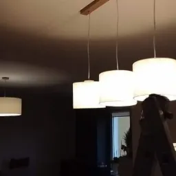 Montaż białych lamp wiszących z abażurami, widoczna aluminiowa drabina w pokoju z choinką w tle.