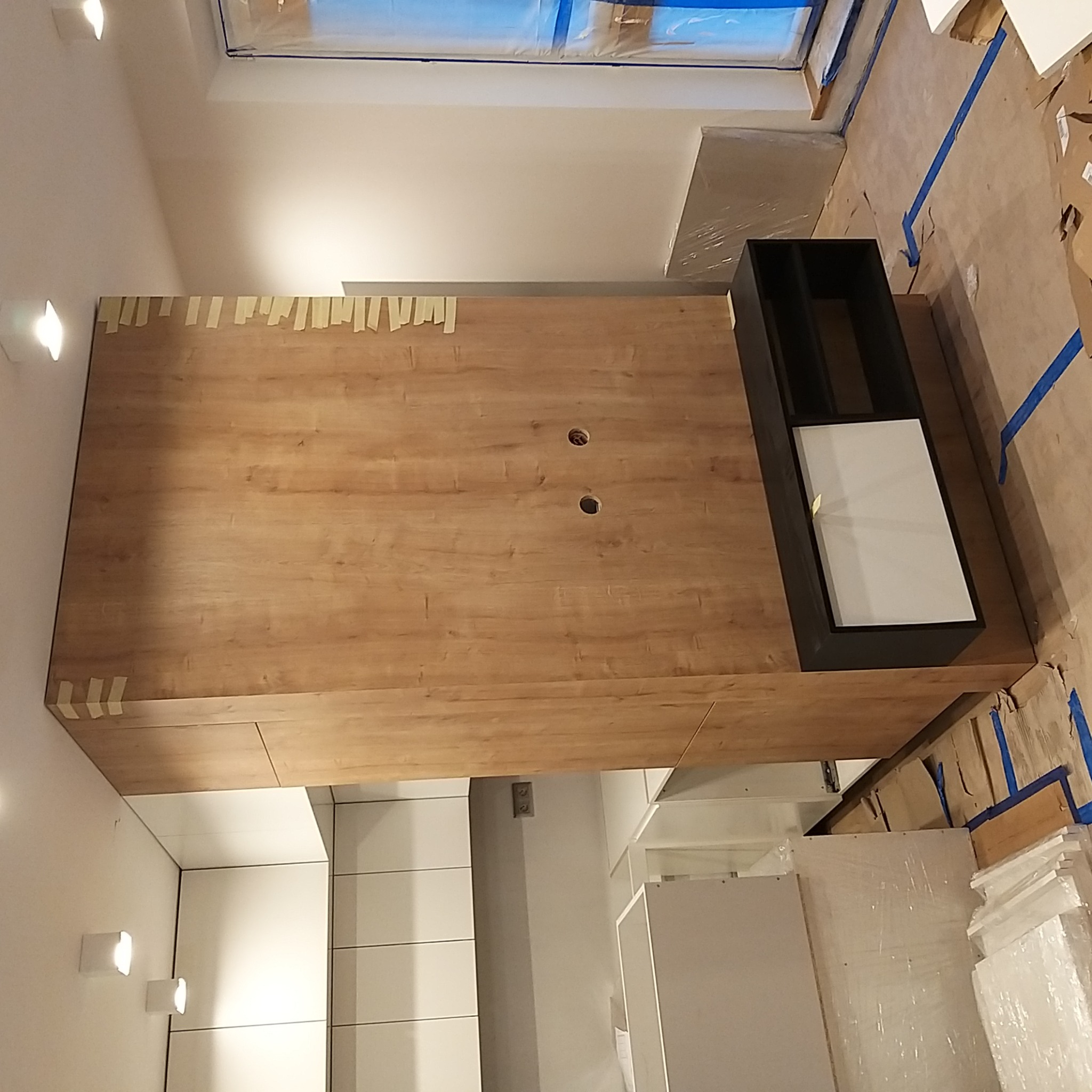 Montaż drewnianej zabudowy meblowej z czarnym wkładem w otworach na kable, zabezpieczonej taśmą malarską w trakcie prac wykończeniowych w nowym mieszkaniu.