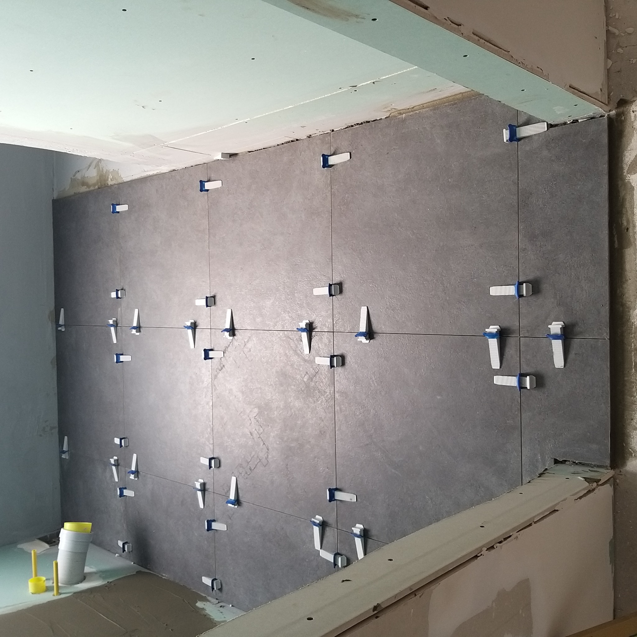 Szare płytki ścienne układane z użyciem systemu poziomowania płytek, widoczne sznurki i klipsy dystansowe, w tle wiaderko i narzędzia do kleju