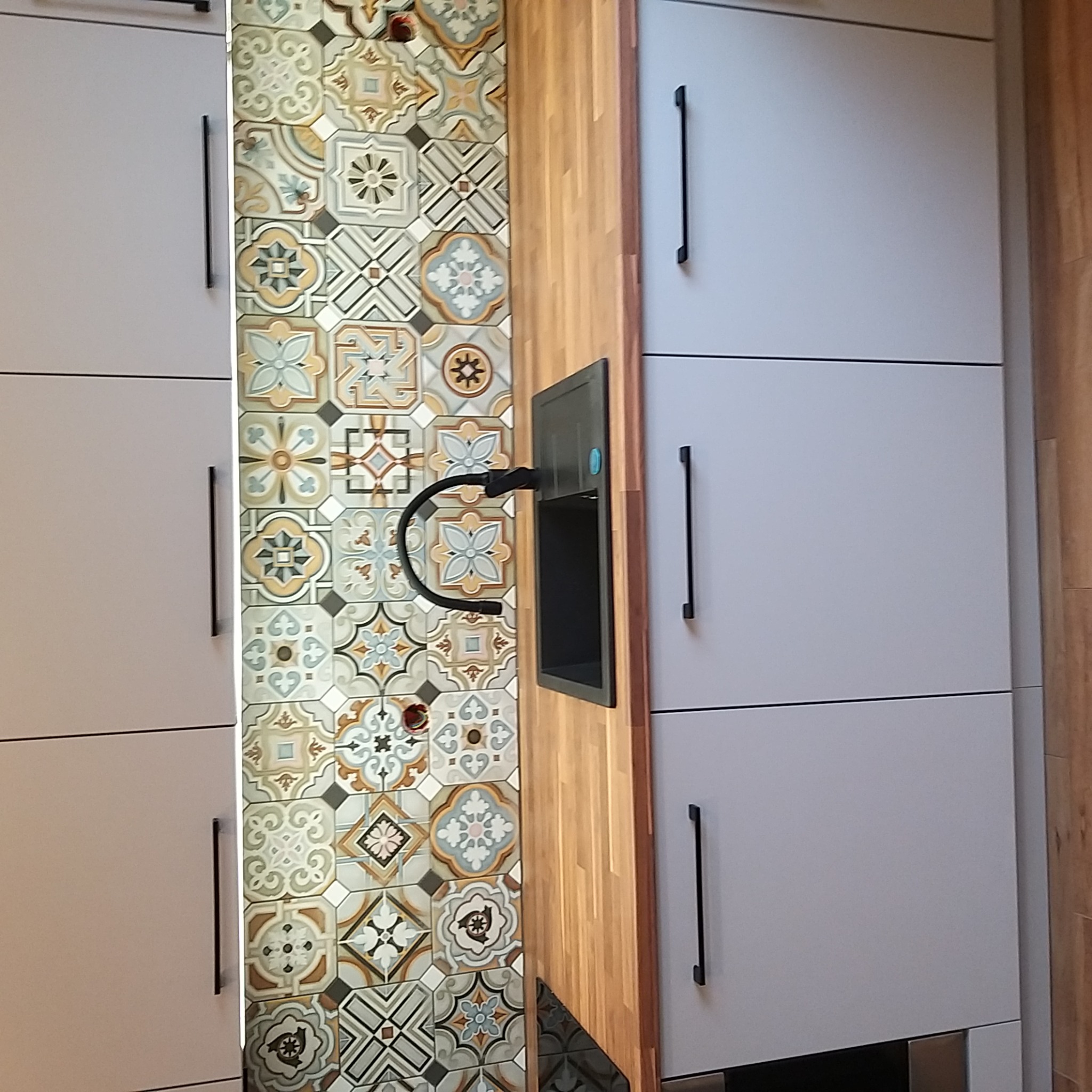 Szare szafki kuchenne z długimi czarnymi uchwytami, drewniany blat i ściana wyłożona kolorowymi, wzorzystymi płytkami ceramicznymi, z widocznym czarnym kranem.