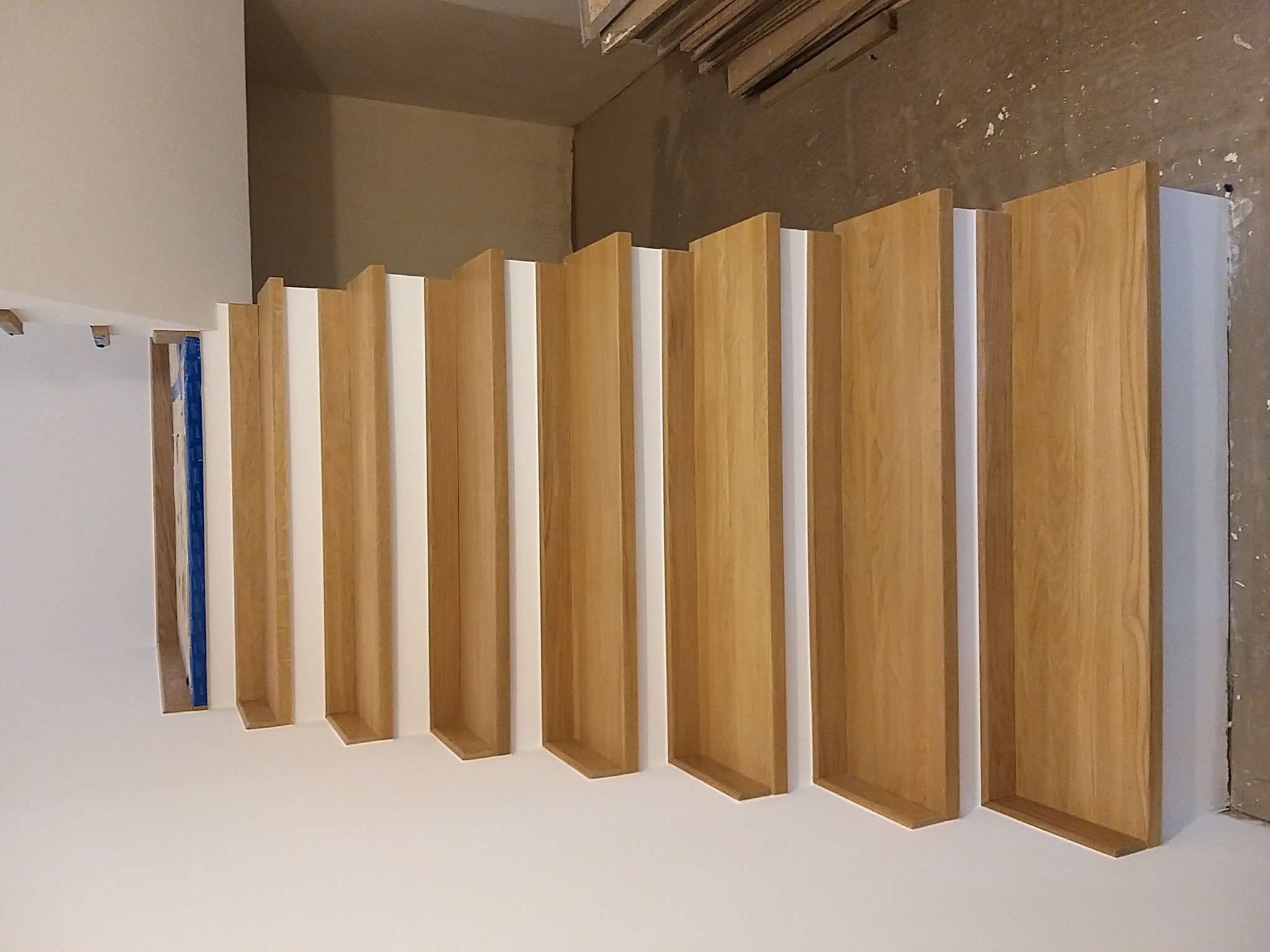 Drewniane panele dekoracyjne na białej ścianie, widok pod kątem z góry, fragment zabezpieczonego folią otworu drzwiowego w tle.