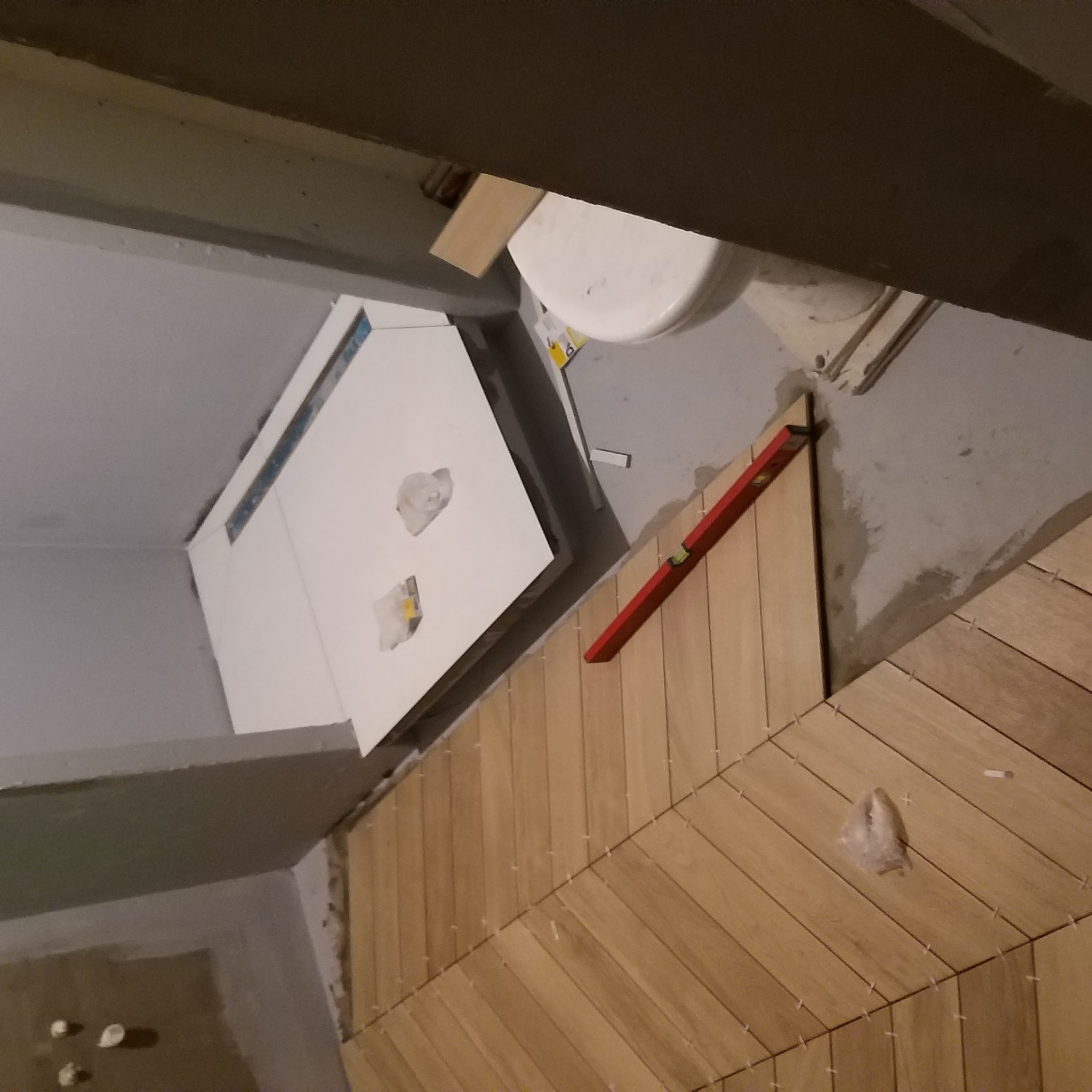 Układanie drewnopodobnych paneli podłogowych w łazience, widoczny poziomica, biała toaleta i przygotowane miejsce pod zabudowę geberit.