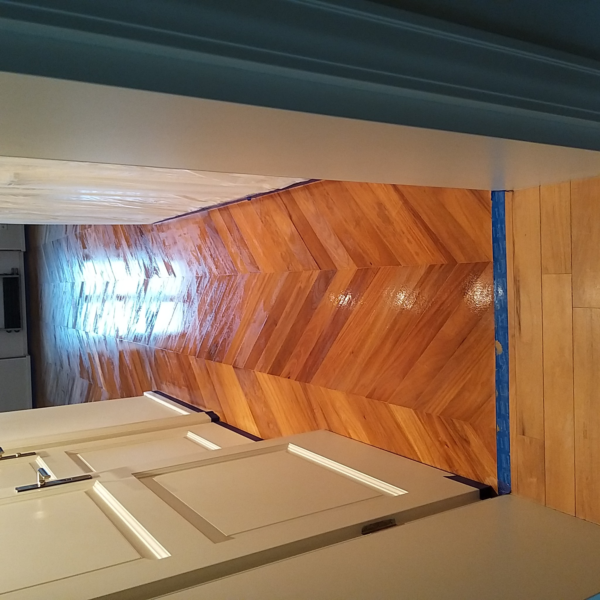 Parkiet ułożony w jodełkę, zabezpieczony niebieską taśmą malarską przy ścianie, widoczne fragmenty białych drzwi.