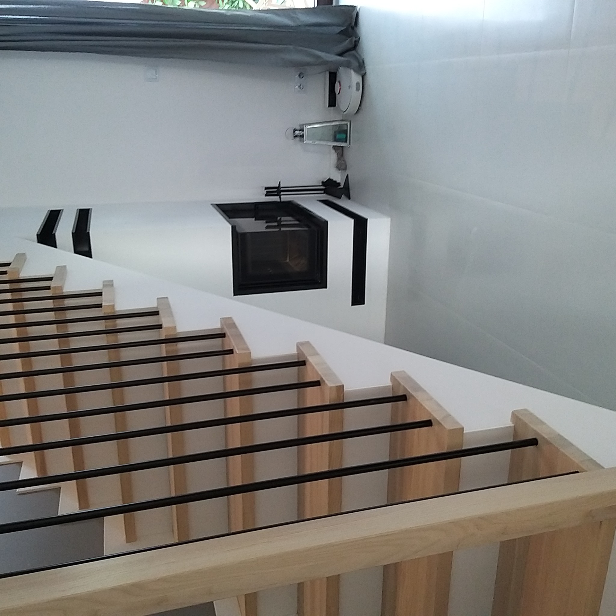 Nowoczesny kominek w obudowie z białego kamienia z czarnymi akcentami, widoczny zza drewnianej konstrukcji schodów z czarnymi poręczami, na ścianie biały robot sprzątający i sterownik.