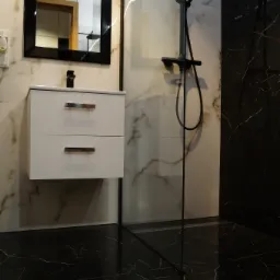 Nowoczesna łazienka z czarnymi marmurowymi płytkami podłogowymi, białą szafką pod umywalką z czarną armaturą, lustrem w czarnej ramie i kabiną prysznicową z czarnym zestawem natryskowym.