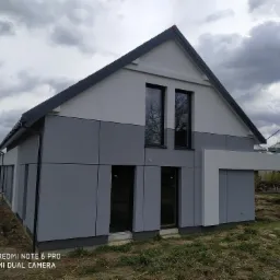 Nowoczesny dom jednorodzinny z elewacją w odcieniach szarości i bieli, widoczny fragment ogrodu i pochmurne niebo.