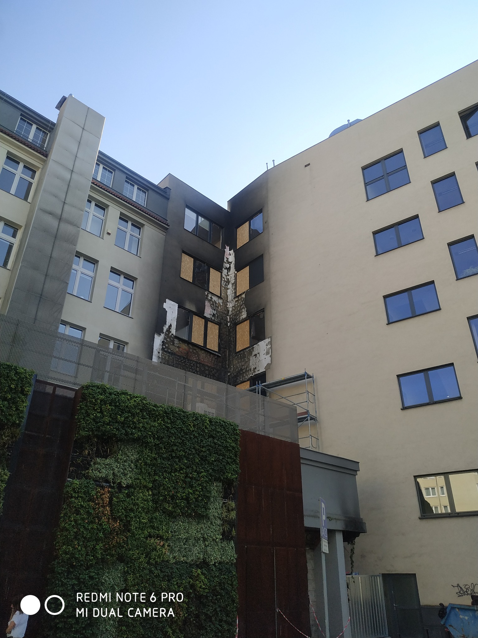 Uszkodzona elewacja budynku po pożarze, okna zabite płytami, widoczne ślady osmolenia i zniszczeń, rusztowanie przy ścianie, fragment zielonej ściany i tablica informacyjna w tle.