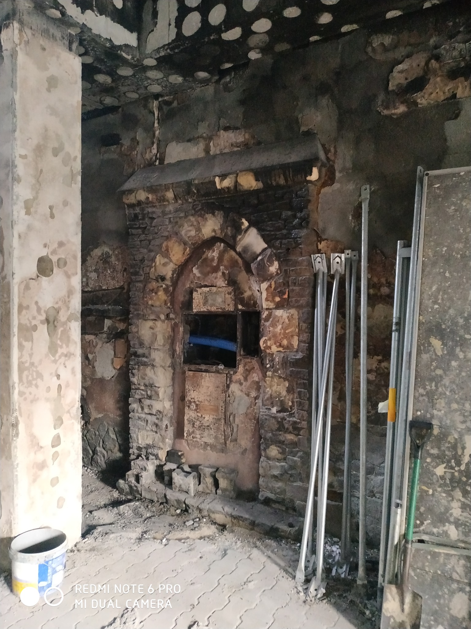 Wnętrze po pożarze z widocznymi uszkodzeniami ścian, ceglanym łukiem nad otworem i elementami rusztowania oraz wiadrem farby.