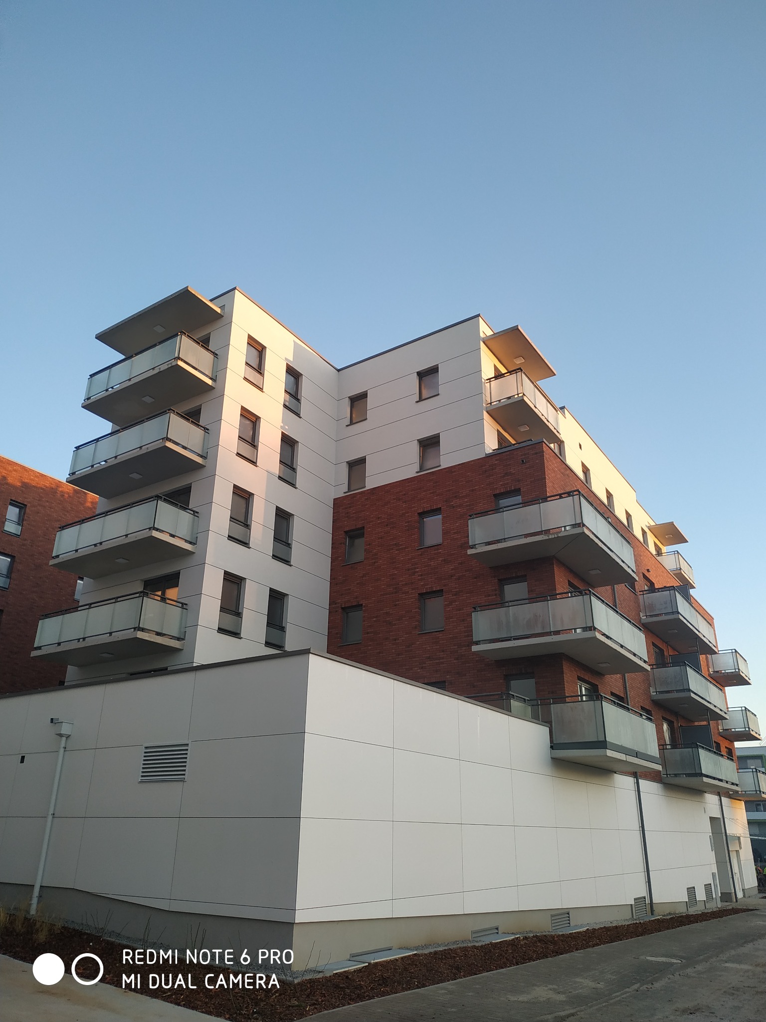 Nowoczesny budynek mieszkalny z balkonami, fragment elewacji z białymi panelami i ceglaną ścianą, widok z dołu w pogodny dzień.