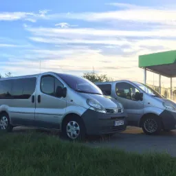 Dwa srebrne busy Opel Vivaro zaparkowane obok myjni samochodowej z zielonym dachem i logo M, na tle błękitnego nieba z delikatnymi obłokami.