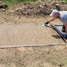 Pracownik w czarnych rękawicach wyrównuje szpachelką nawierzchnię z drobnych kamyczków w beżowym odcieniu, ograniczoną szarą kostką, z widoczną linią podziału na obszar z szarym grysem. Na drugim...
