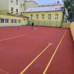 Malowanie linii na czerwonym boisku sportowym z użyciem specjalistycznego wózka malarskiego, budynek szkoły w tle.