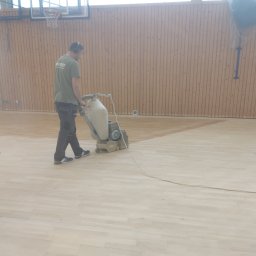 Michał Dobija - Pracownik w słuchawkach szlifuje parkiet w sali gimnastycznej za pomocą specjalistycznej maszyny, widoczna różnica w odcieniu parkietu po obróbce.