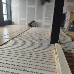 Michał Dobija - Układanie paneli podłogowych w Łodygowicach: widok na podkład z listew, częściowo pokryty panelami, pracownik w tle aplikuje klej przy ścianie, widoczne okno i czarna kolumna.