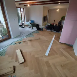 Proces układania parkietu w jodełkę francuską, widoczne fragmenty ułożonej podłogi, przygotowane deski, narzędzia i pracownik szlifujący podłoże w pomieszczeniu w trakcie remontu.