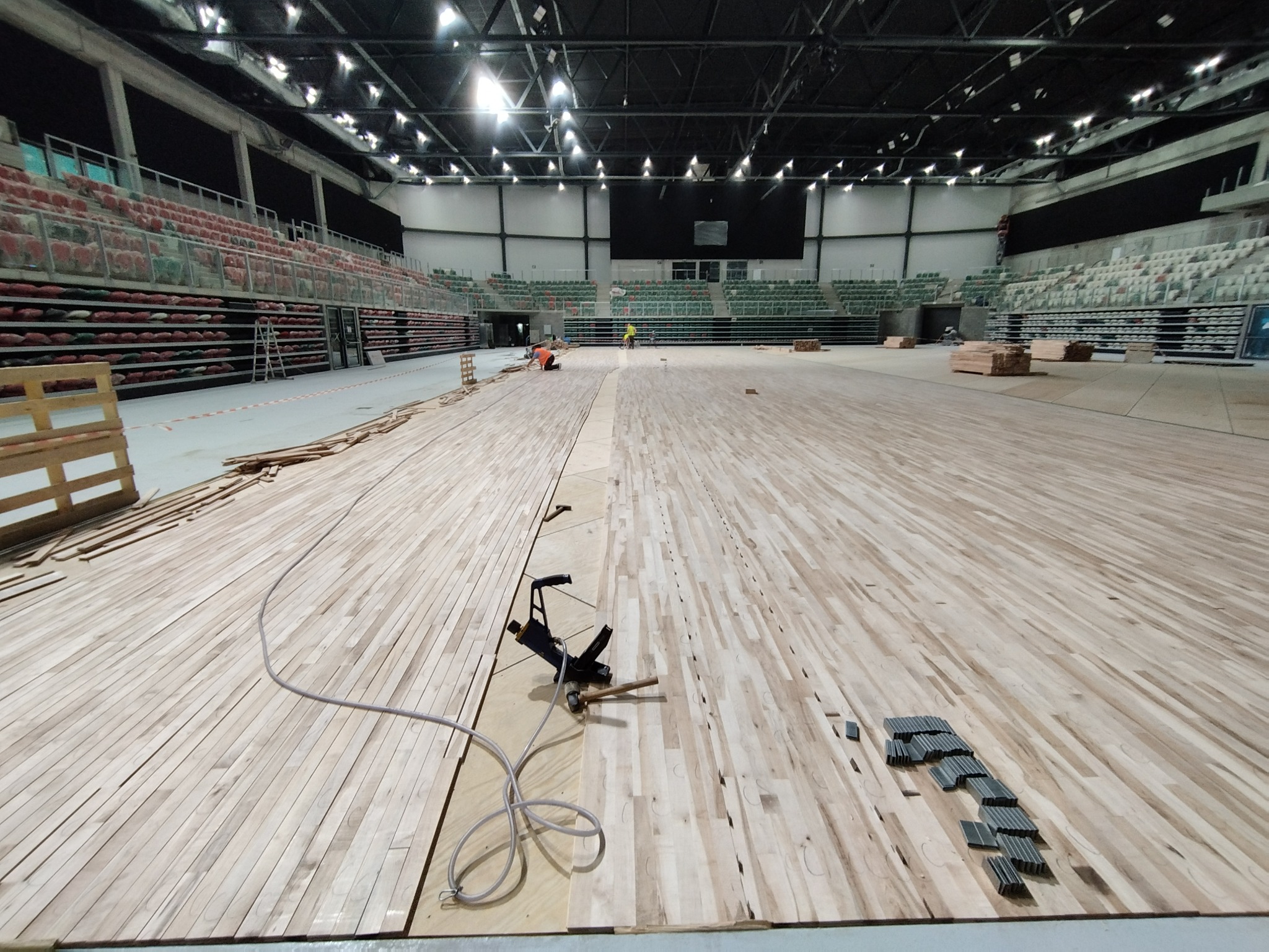 Duża hala sportowa w trakcie renowacji podłogi; widoczne jasne deski parkietowe, narzędzia do montażu i pracownicy w oddali.