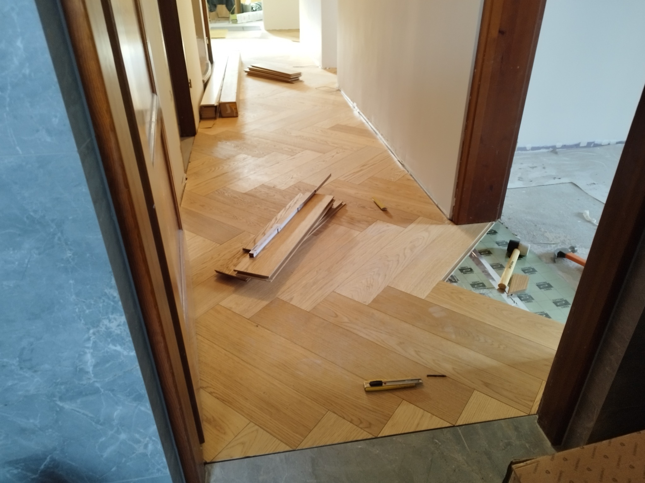 Fragment podłogi w trakcie układania parkietu w jodełkę; widoczne narzędzia, młotek, nożyk oraz paczka desek podłogowych.