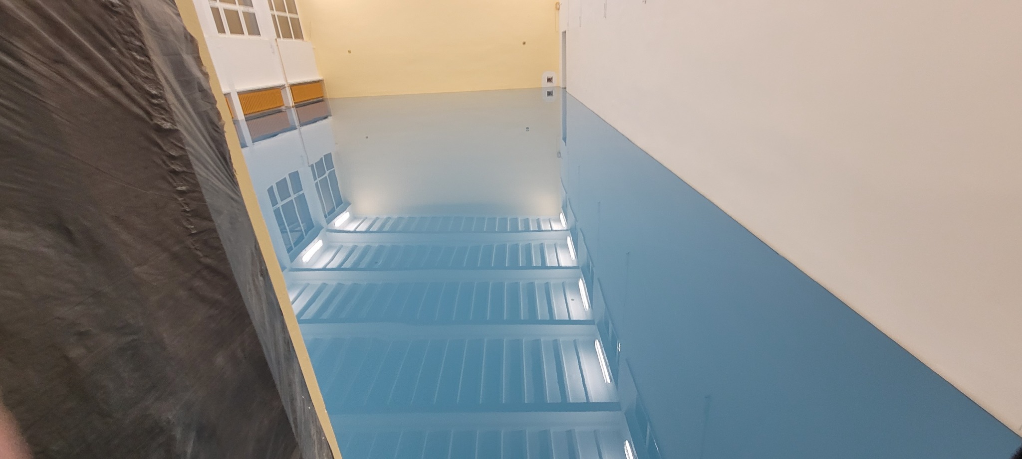 Świeżo pomalowana, błyszcząca na niebiesko ściana w dużym pomieszczeniu biurowym, z widocznym fragmentem zabezpieczonej folią podłogi i odbiciem okien w podłodze.