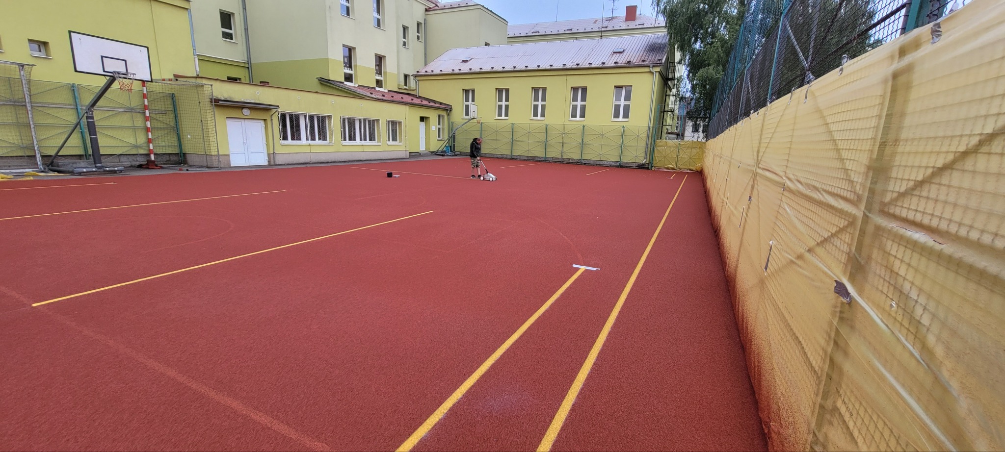 Malowanie linii na czerwonym boisku sportowym z użyciem specjalistycznego wózka malarskiego, budynek szkoły w tle.