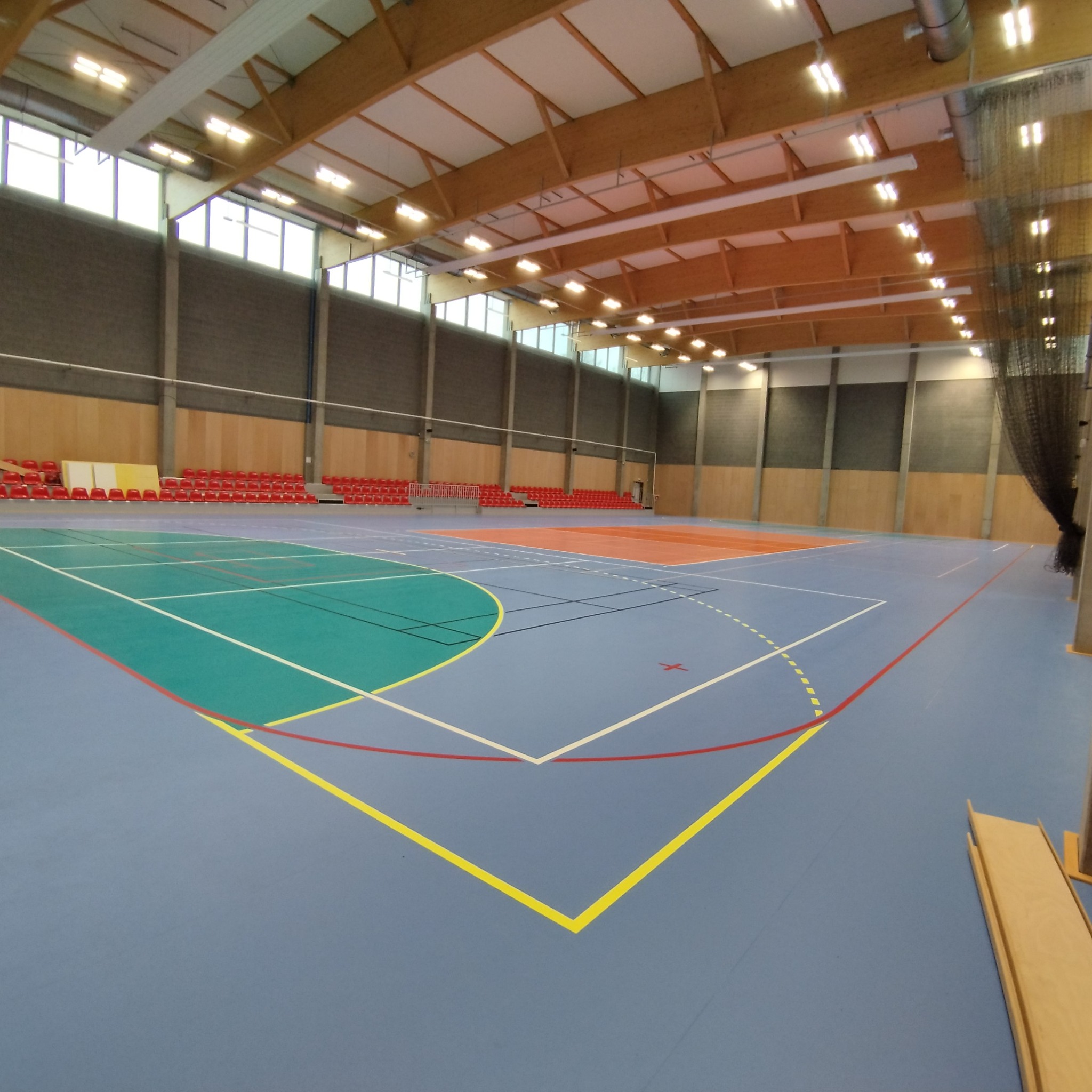 Wnętrze dużej hali sportowej z parkietem w kolorze niebieskim, zielonym i pomarańczowym, widoczne linie boisk, trybuny z czerwonymi krzesłami w tle oraz drewniana konstrukcja dachu z oświetleniem.