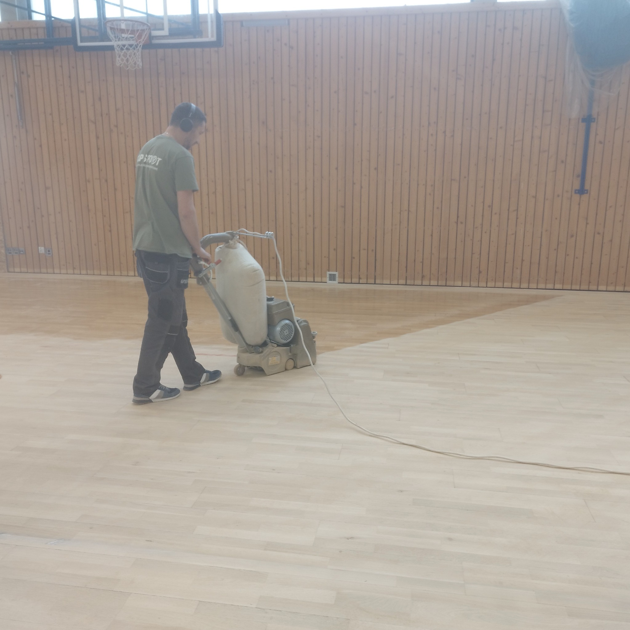Pracownik w słuchawkach szlifuje parkiet w sali gimnastycznej za pomocą specjalistycznej maszyny, widoczna różnica w odcieniu parkietu po obróbce.